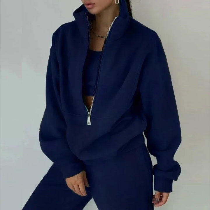 Une femme porte un ensemble décontracté bleu marine avec un haut à zip et un pantalon assorti. Le haut est légèrement oversize avec un col zippé, créant un look moderne et confortable. Le fond est neutre et minimaliste.